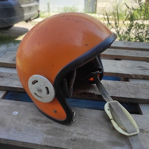 May include: A bright orange open-face motorcycle helmet featuring a black trim along the edge, a white circular side attachment, and a grey chin strap with a white plastic buckle. The helmet has a smooth, glossy finish with minor surface wear.
