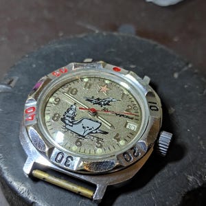 May include: A silver-tone analog wristwatch with a textured gold-colored dial featuring a graphic of a ship and a jet. The watch face includes a red star, Arabic numerals, a date window, and a rotating bezel with red and black markings.