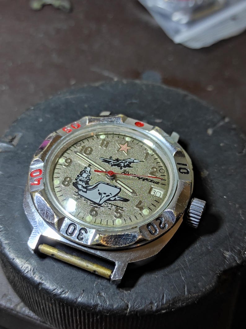 May include: A silver-tone analog wristwatch with a textured gold-colored dial featuring a graphic of a ship and a jet. The watch face includes a red star, Arabic numerals, a date window, and a rotating bezel with red and black markings.