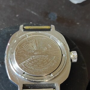 May include: A silver-toned metal wristwatch case with a circular back featuring an engraved design of a sun rising over water. The case includes a side crown and two spring bars for a strap. Cyrillic text is embossed along the bottom edge.