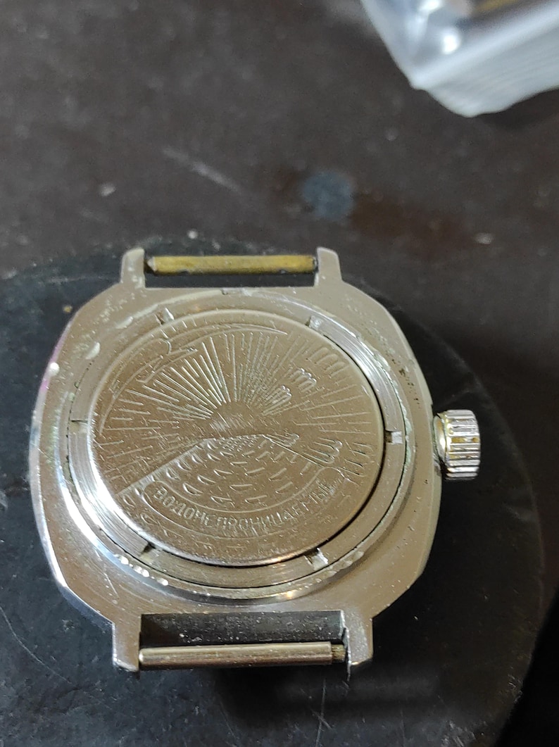May include: A silver-toned metal wristwatch case with a circular back featuring an engraved design of a sun rising over water. The case includes a side crown and two spring bars for a strap. Cyrillic text is embossed along the bottom edge.