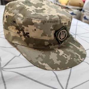 May include: A military-style patrol cap featuring a digital camouflage pattern in shades of olive green, tan, and brown. The cap has a flat top, a stiff brim, and a small, square-shaped patch on the front displaying the Ukrainian trident emblem.