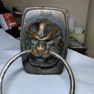 Könnte beinhalten: Ein Türklopfer in Bronzefarbe mit einem detaillierten Löwenkopf und einem silberfarbenen Ring. Das Gesicht des Löwen weist aufwendige Details auf und ist auf einer rechteckigen Platte montiert. Das Design hat eine antike oder Vintage-Ästhetik.