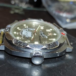 May include: A silver-tone wristwatch with a metallic gold dial featuring a graphic of a submarine and a red star. The watch has a rotating bezel with black engraved numbers, a circular crown on the side, and a clear crystal covering the face.