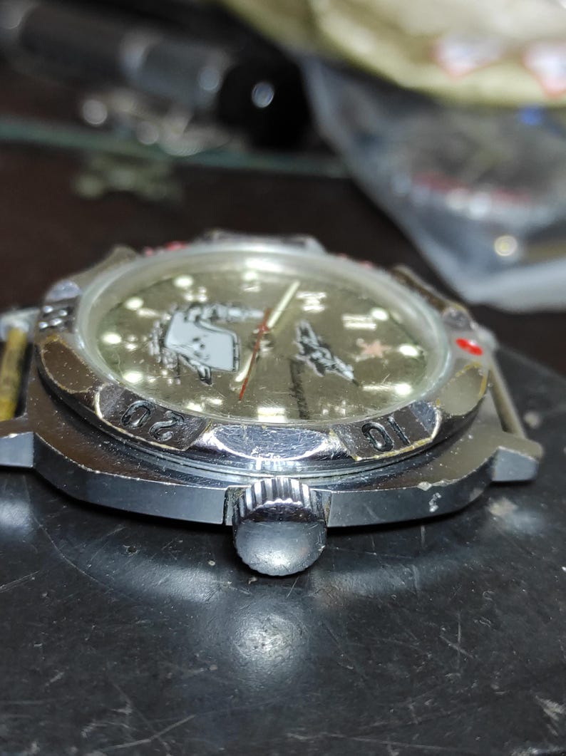 May include: A silver-tone wristwatch with a metallic gold dial featuring a graphic of a submarine and a red star. The watch has a rotating bezel with black engraved numbers, a circular crown on the side, and a clear crystal covering the face.