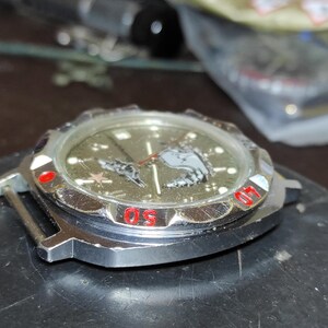 May include: A silver-toned wristwatch case with a metallic olive green dial featuring a graphic of a submarine and a red star. The rotating bezel has red numerical markings for 40 and 50, and the watch face includes silver hour markers and hands.