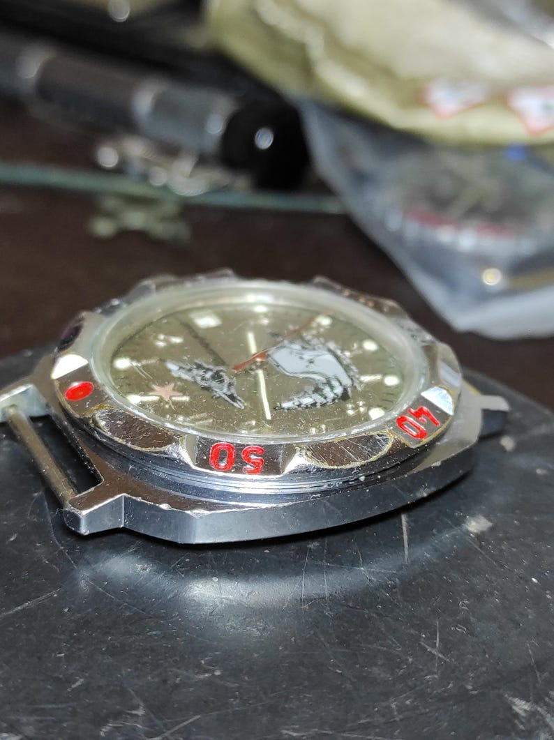 May include: A silver-toned wristwatch case with a metallic olive green dial featuring a graphic of a submarine and a red star. The rotating bezel has red numerical markings for 40 and 50, and the watch face includes silver hour markers and hands.