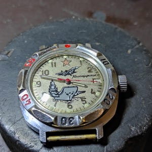 May include: A silver-tone wristwatch with a textured beige dial featuring a red star, a black airplane graphic, and a ship illustration. The dial includes the Cyrillic text "Командирские". It has a rotating bezel with red and black numerals.