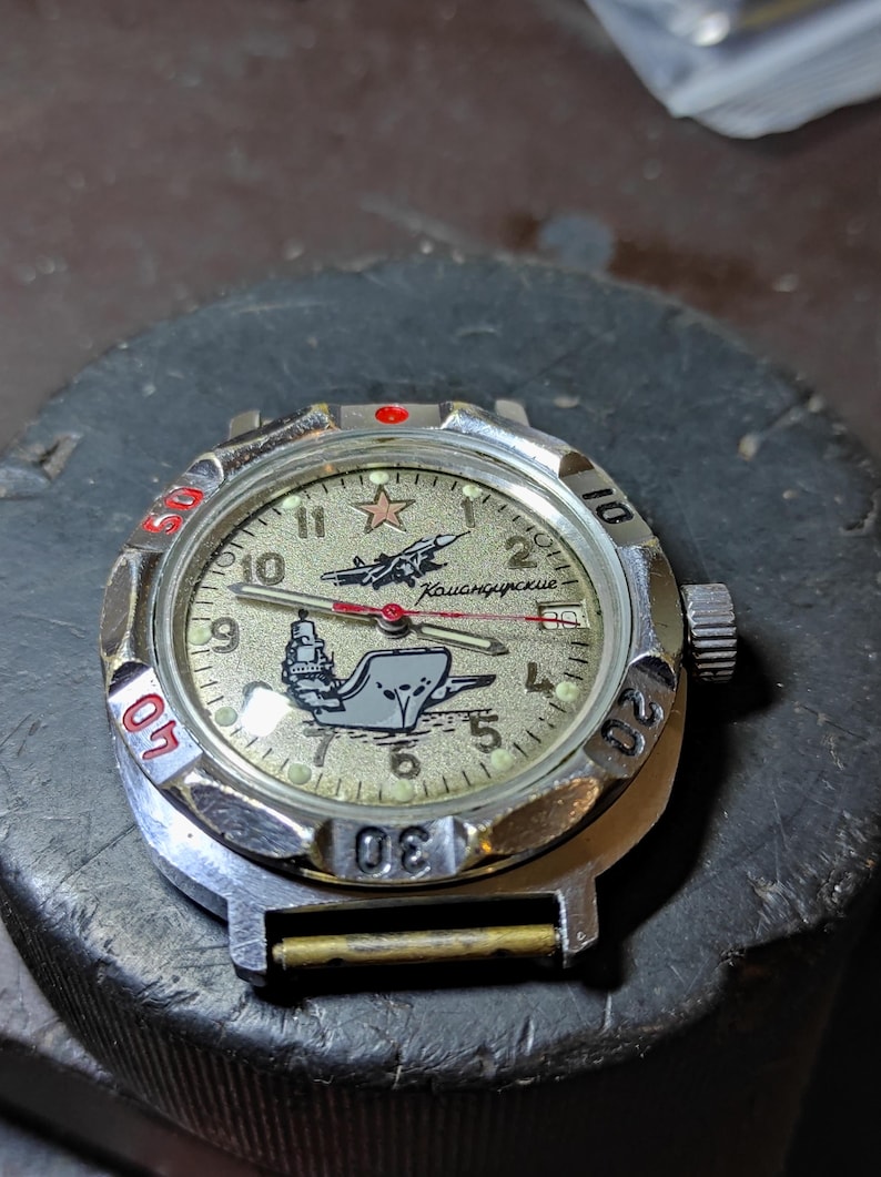 May include: A silver-tone wristwatch with a textured beige dial featuring a red star, a black airplane graphic, and a ship illustration. The dial includes the Cyrillic text "Командирские". It has a rotating bezel with red and black numerals.