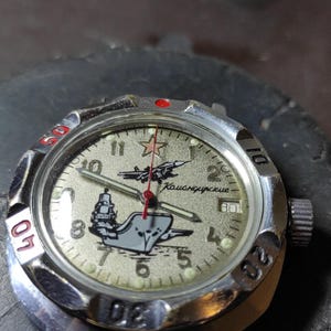 May include: A silver-tone analog wristwatch with a textured beige dial featuring a red star, a jet plane, and a ship graphic. The dial includes the Cyrillic text "Командирские" and Arabic numerals. It has a rotating bezel with red and black markings.