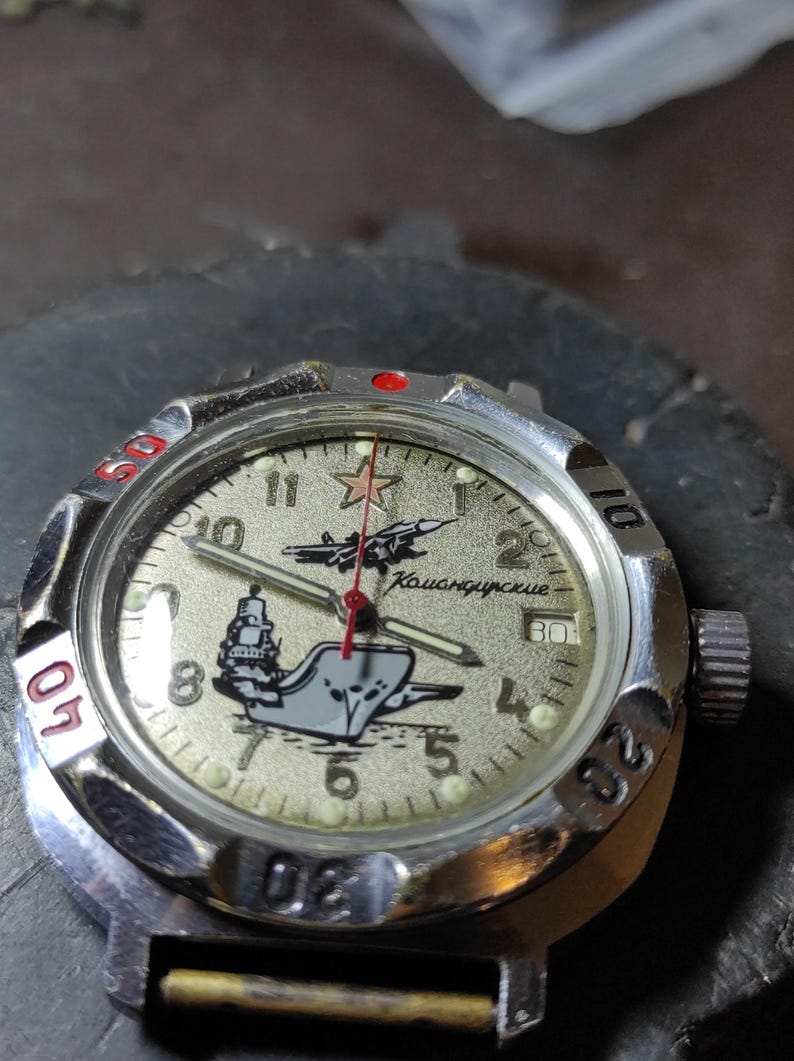 May include: A silver-tone analog wristwatch with a textured beige dial featuring a red star, a jet plane, and a ship graphic. The dial includes the Cyrillic text "Командирские" and Arabic numerals. It has a rotating bezel with red and black markings.