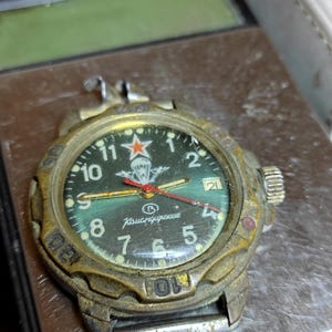 Puede incluir: Reloj de pulsera vintage con esfera verde, números blancos y detalles rojos. El reloj tiene un aspecto metálico envejecido, con el texto "Komandirskie" visible. La esfera presenta una estrella roja y un emblema de paracaídas.