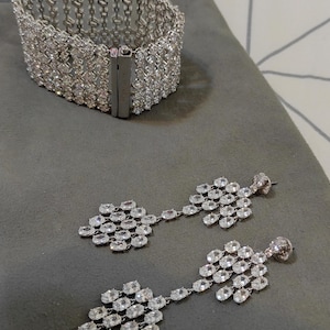 May include: A silver-toned cuff bracelet and a pair of matching drop earrings, each featuring a multi-row lattice design encrusted with numerous clear, oval-cut stones. The bracelet includes a rectangular metal clasp closure.