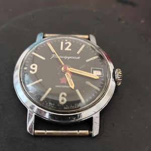 May include: A vintage mechanical wristwatch with a round silver-tone case and a black dial. The dial features white Arabic numerals at 6, 9, and 12, a red star, gold-tone hands, a date window at the 3 o'clock position, and Cyrillic text.