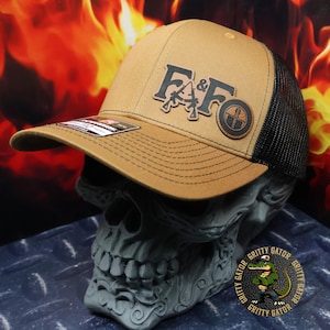 May include: A tan and black trucker hat with a brown leather patch that says "F&F" with two rifles crossed underneath. The patch is on the front of the hat. The hat has a black mesh back.