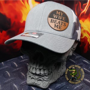 May include: Gray baseball cap with a leather patch that reads "MY WIFE BEATS ME." The cap has a mesh back with a star pattern and a curved brim. The hat is displayed on a skull-shaped stand.