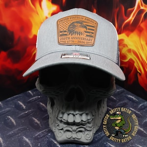 May include: A gray trucker hat with a leather patch featuring an eagle, American flag, and text reading "UNITED STATES OF AMERICA" and "250TH ANNIVERSARY 1776-2026." The hat has a white mesh back and a curved brim. The hat is displayed on a gray skull.