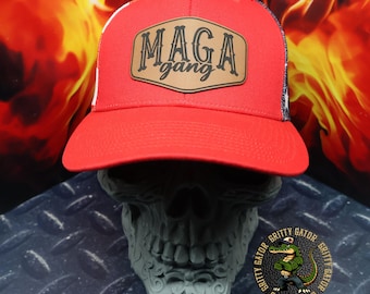 May include: Red and white trucker hat with a brown leather patch that says "MAGA gang". The hat is sitting on a gray skull with a black and white alligator logo.