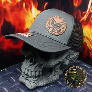May include: Gray trucker hat with a black mesh back. The hat features a brown leather patch with the text "IN GOD WE TRUST" and "THE GUNS ARE JUST BACKUP" surrounding an eagle emblem. The hat is displayed on a skull.