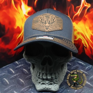 May include: A dark blue trucker hat with a brown leather patch featuring a stylized deer skull design. The patch has a wood grain texture and the hat has a mesh back.
