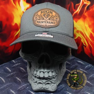 May include: A gray trucker hat with a black mesh back and a curved brim. The hat features a brown leather patch with the words "HIGH MAINTENANCE" and a diamond design. The hat is displayed on a gray skull.