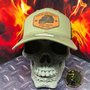 May include: Olive green trucker hat with a black mesh back. The hat features a brown leather patch with the text "TREAD ON THOSE WHO TREAD ON YOU" and a graphic of a bulldozer. The hat is displayed on a gray skull.