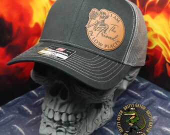 May include: A charcoal gray trucker hat with a gray mesh back. A circular leather patch features a skeleton holding a beer and the text "I AM The Friend IN LOW PLACES". The hat is displayed on a skull-shaped stand.