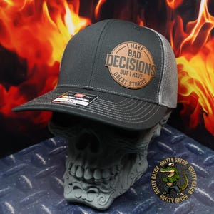 May include: A black and gray trucker hat with a brown leather patch that says "I make bad decisions but I have great stories".