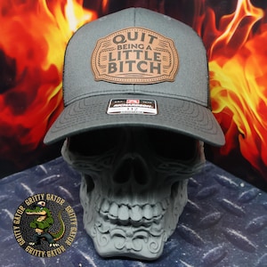 May include: A gray trucker hat with a black mesh back and a brown leather patch. The patch features the text "QUIT BEING A LITTLE BITCH" in a bold font. The hat is displayed on a gray skull.
