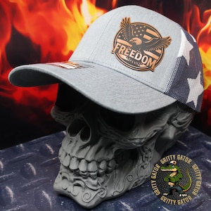 May include: A light gray baseball cap with a mesh back featuring white stars. The front of the cap has a brown leather patch with an eagle, American flag design, and the word "FREEDOM". The cap is displayed on a gray skull.