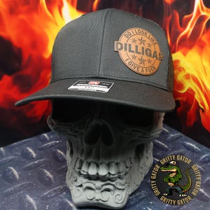 May include: A black trucker hat with a curved brim and mesh back. The hat features a brown leather patch with the text "DO I LOOK LIKE DILLIGAS I GIVE A FUCK". The hat is displayed on a gray skull.