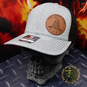 May include: A gray and black trucker hat with a brown leather patch that says "Don't Tread On Me" and "F.A.F.O.". The patch features a coiled rattlesnake.