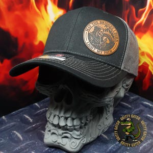 May include: A black and gray trucker hat with a curved brim and mesh back. The hat features a circular patch with the text "MAY THE BRIDGES I BURN LIGHT MY PATH" and an illustration of a Grim Reaper figure.