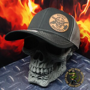 May include: A dark gray Oilfield Roughneck trucker hat with a mesh back and a curved brim. The hat features a circular leather patch with a skull wearing a hard hat and crossed tools. The background is a fiery orange and yellow.