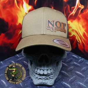 May include: A tan and black trucker hat with the phrase "NOT my problem" in a copper-colored patch. The hat is displayed on a gray skull statue. The background features flames and the Gritty Gator logo.