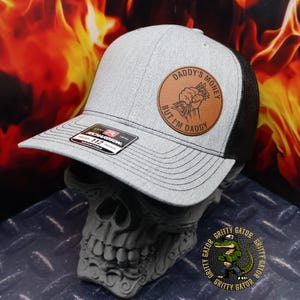 May include: A heather gray and black trucker hat featuring a round brown leather patch on the front. The patch displays a graphic of a fist holding cash with the text DADDY'S MONEY BUT I'M DADDY. A black sticker is on the brim with Richardson 112 branding.