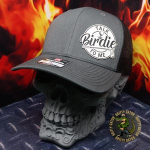 May include: A gray baseball cap with a black mesh back. The cap features a white circular patch with the words "Talk Birdie To Me" and a golf ball and club graphic. The cap is displayed on a skull-shaped stand.