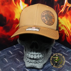 Puede incluir: Gorra de camionero marrón y negra con las palabras "Liberty or Death" impresas en un parche circular. El sombrero se exhibe sobre un objeto gris con forma de calavera. La gorra tiene una visera curva y una parte trasera de malla.