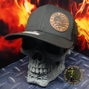 May include: A black baseball cap with a mesh back and a curved brim. The cap features a circular leather patch with the text "ONE NATION UNDER GOD" and a patriotic design. The cap is displayed on a gray skull-shaped stand.