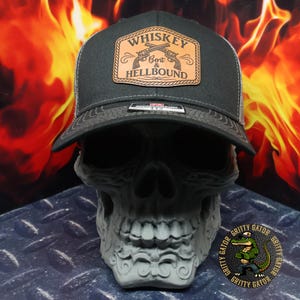 May include: A black trucker hat with a leather patch that reads "Whiskey Bent & Hellbound" with crossed pistols. The hat has a gray mesh back and is displayed on a gray skull.