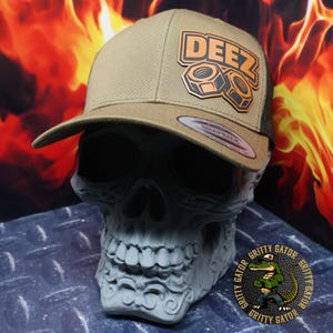 May include: A tan and black trucker hat with the word "DEEZ" and two nuts in a brown patch on the front. The hat has a curved brim and a mesh back. The hat is displayed on a gray skull.