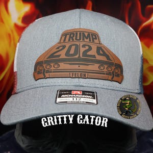 May include: Gray trucker hat with a brown leather patch featuring the text "TRUMP 2024" and a car design. The hat has a white mesh back and the words "GRITTY GATOR" are printed on the front.