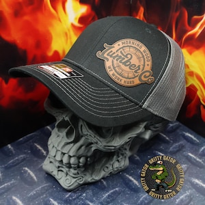 May include: A black and gray trucker hat with a brown leather patch that says "Morning Wood Lumber Co. We Work Hard".