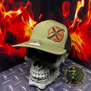 May include: Olive green and black trucker hat with a leather patch that reads "generation X 1965-1980". The hat is displayed on a skull-shaped decorative object. The background features a fiery design.