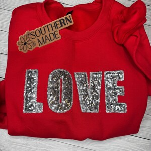 May include: A bright red sweatshirt with the word "LOVE" spelled out in silver sequins. A wooden tag with the words "SOUTHERN MADE" is attached to the neckline. The sweatshirt is folded.
