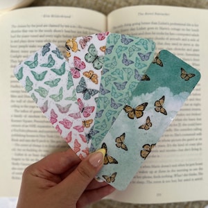 May include: Four colorful bookmarks with a butterfly pattern. The bookmarks are made of paper and have a watercolor effect. The butterflies are in shades of pink, blue, green, and yellow.
