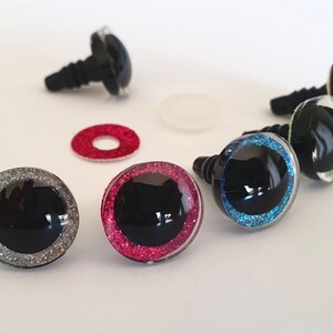 Puede incluir: Un conjunto de ojos de seguridad de pl&aacute;stico para manualidades, con centros negros rodeados por anillos brillantes en plata, rosa, azul y verde. Cada ojo tiene un poste de pl&aacute;stico roscado e incluye una arandela circular separada para su fijaci&oacute;n.