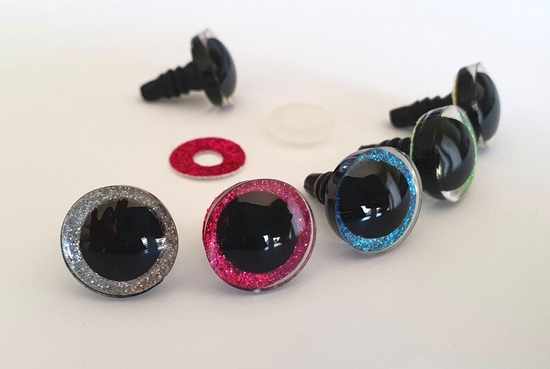 Puede incluir: Un conjunto de ojos de seguridad de pl&aacute;stico para manualidades, con centros negros rodeados por anillos brillantes en plata, rosa, azul y verde. Cada ojo tiene un poste de pl&aacute;stico roscado e incluye una arandela circular separada para su fijaci&oacute;n.