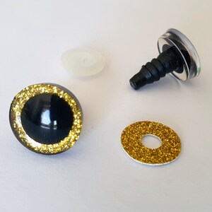 Puede incluir: Un juego de ojos para manualidades con un domo negro brillante rodeado por un anillo de purpurina dorada. El conjunto incluye un poste de pl&aacute;stico negro con una arandela transparente y una arandela de purpurina dorada para su fijaci&oacute;n.
