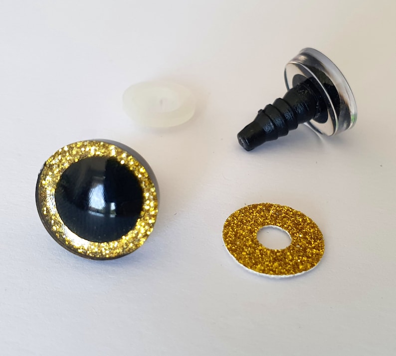 Puede incluir: Un juego de ojos para manualidades con un domo negro brillante rodeado por un anillo de purpurina dorada. El conjunto incluye un poste de pl&aacute;stico negro con una arandela transparente y una arandela de purpurina dorada para su fijaci&oacute;n.